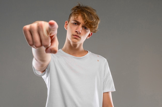 Hübscher emotionaler Teenagerjunge, der im Studio gegen grau, Kerl in einem grauen T-Shirt aufwirft, zeigt einen Finger an der Kamera