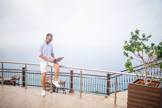 Hübscher erfolgreicher junger männlicher Geschäftsmann, der mit Laptop arbeitet, betrachtet das Mittelmeer. Er trägt ein Hemd und weiße Shorts. Fernarbeit im Urlaub. Urlaubskonzept