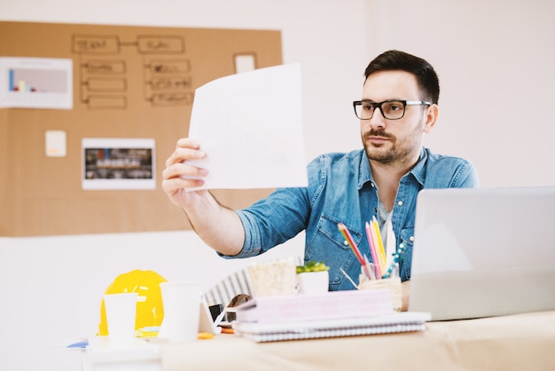 Hübscher fokussierter junger Designer, der Briefing-Papier beim Sitzen am Schreibtisch betrachtet.