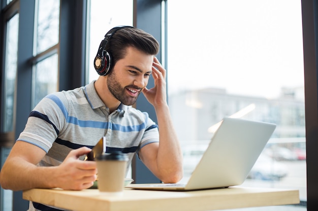 Hübscher freudiger glücklicher mann, der kopfhörer trägt und musik hört, während er den laptopbildschirm betrachtet