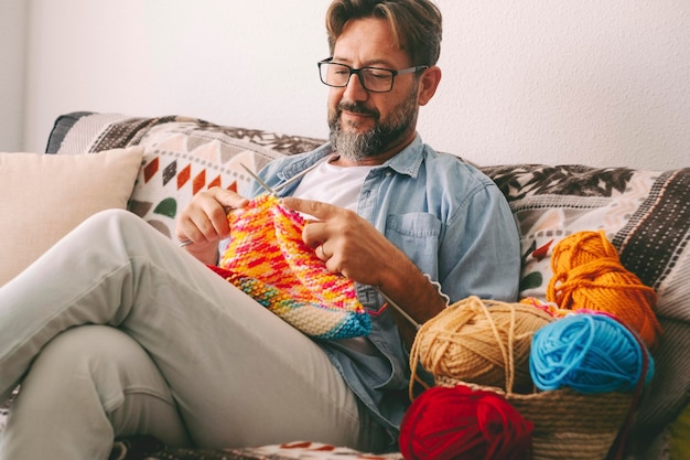 Hübscher, fröhlicher erwachsener Mann, der zu Hause mit Wolle und Nadeln strickt. Männliche Menschen bei weiblichen üblichen Aktivitäten. Entspannung und Hobby allein bei Indoor-Freizeitaktivitäten. Ältere Menschen sitzen auf dem Sofa