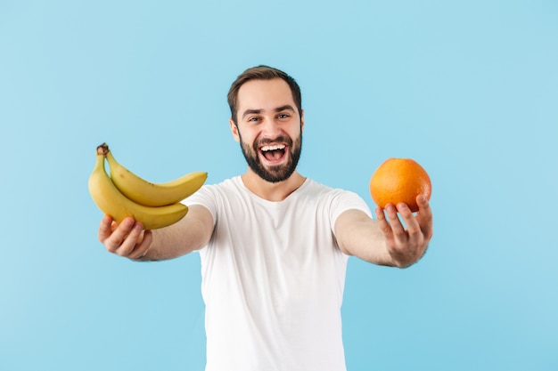 Hübscher fröhlicher junger bärtiger Mann mit T-Shirt, der isoliert über blauer Wand steht und Orange und Banane zeigt