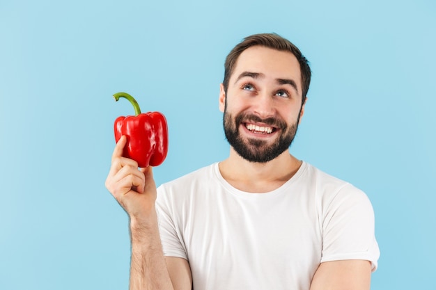 Hübscher fröhlicher junger bärtiger Mann mit T-Shirt, der isoliert über blauer Wand steht und rote Paprika zeigt