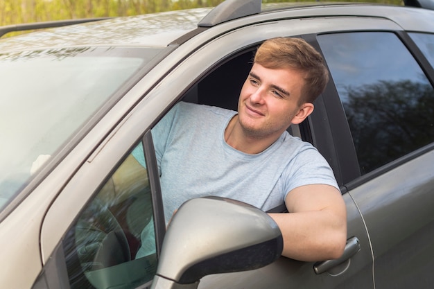 Hübscher fröhlicher Kerl, Fahrer, junger positiver Mann, sein Auto fahrend, lächelnd, ragen vom Autofenster heraus. Glücklicher Käufer eines neuen Autos, das das Fahren genießt. Emotion, Glück, Freude Konzept