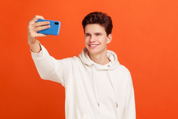 Hübscher, fröhlicher Teenager im lässigen Hoodie mit Smartphone, der Selfie macht und aufrichtig lächelt, Gadget-Sucht. Studioaufnahme im Innenbereich isoliert auf orangefarbenem Hintergrund