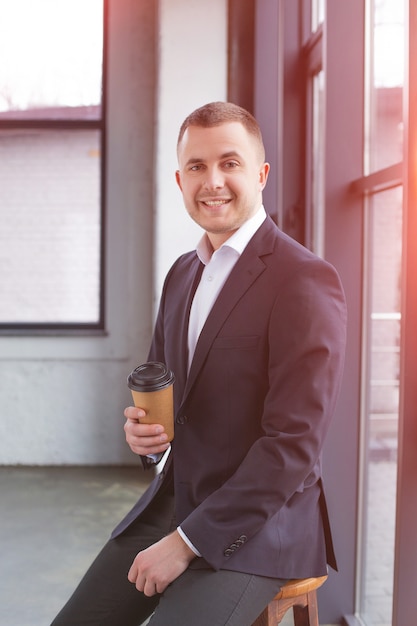 Hübscher Geschäftsmann, der einen schwarzen Anzug mit einer Kaffeetasse trägt