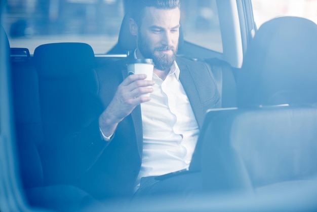 Hübscher Geschäftsmann, der mit Kaffee sitzt, um auf dem Rücksitz des Autos zu gehen. Blick durchs Fenster