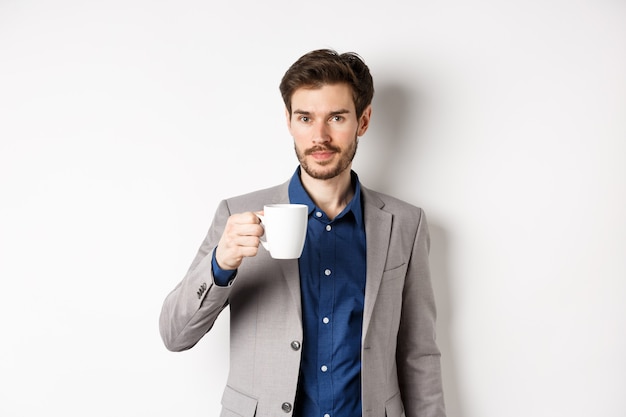 Hübscher Geschäftsmann im Anzug, der Kaffee oder Tee vom Bürobecher trinkt und gegen weißen Hintergrund steht.