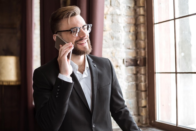 Foto hübscher geschäftsmann in anzug und brille, der im büro am fenster telefoniert