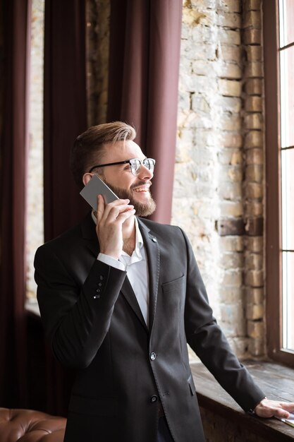 Hübscher Geschäftsmann in Anzug und Brille, der im Büro am Fenster telefoniert