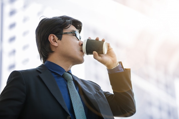 Hübscher Geschäftsmann mit Papierkaffeetasse über Bürogebäude unter dem Sonnenlicht