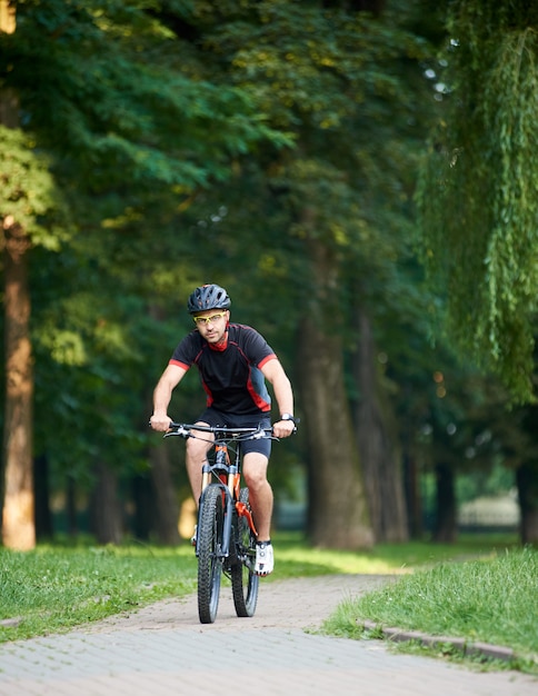 Hübscher getönter Radfahrer in Radsportkleidung, Blick auf die Kamera, während Fahrrad am Sommertag die Parkgasse hinunter fährt