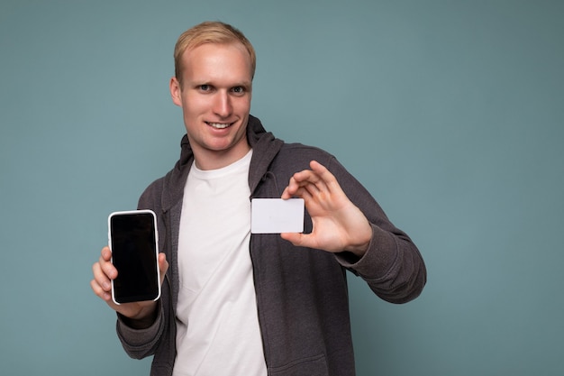 Hübscher glücklich lächelnder blonder mann mit grauem pullover und weißem t-shirt einzeln auf blauer hintergrundwand mit kreditkarte und handy mit leerem bildschirm für mock-up mit blick in die kamera