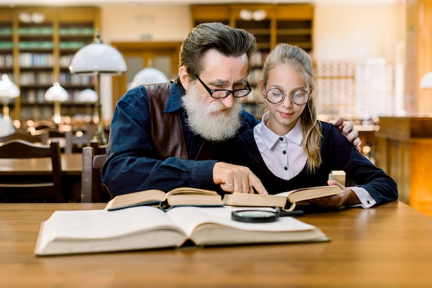 Hübscher Großvater, der ein Buch für seine süße Enkelin liest und sie umarmt, während er zusammen am Tisch in der alten Vintage-Bibliothek sitzt.