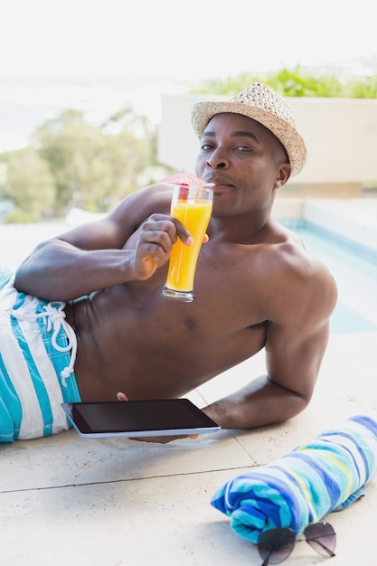 Hübscher hemdloser Mann, der tragendes Cocktail des Tabletten-PC Poolside verwendet