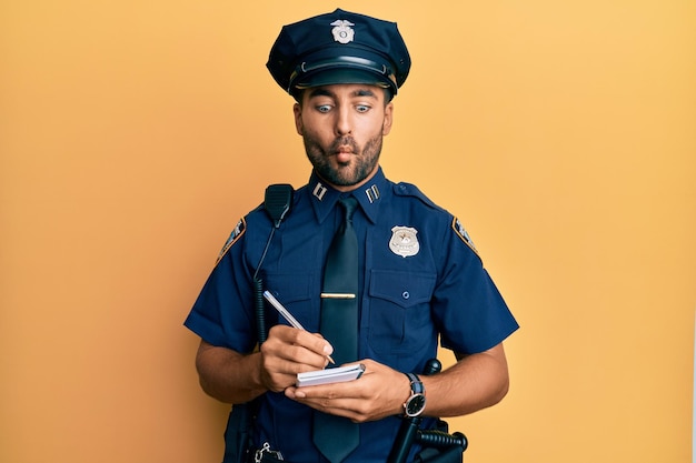 Foto hübscher hispanischer mann in polizeiuniform, der verkehrsstrafe schreibt und ein fischgesicht mit mund und zusammengekniffenen augen verrückt und komisch macht