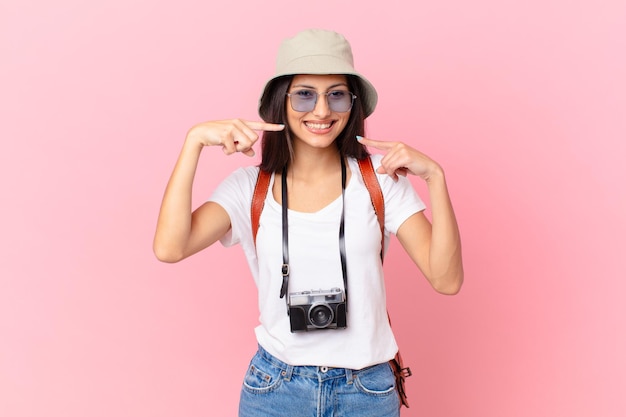 Hübscher hispanischer Tourist, der selbstbewusst auf sein eigenes breites Lächeln mit einer Fotokamera und einem Hut lächelt