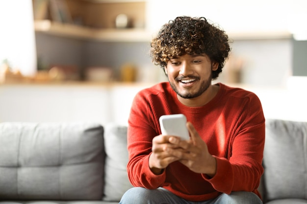 Hübscher Inder, der Smartphone benutzt, während er sich zu Hause auf der Couch entspannt