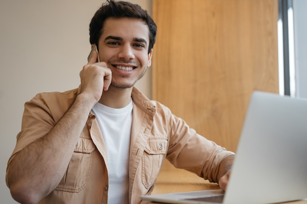 Hübscher indischer Mann, der Laptop verwendet, der auf Handy spricht, das von zu Hause aus arbeitet