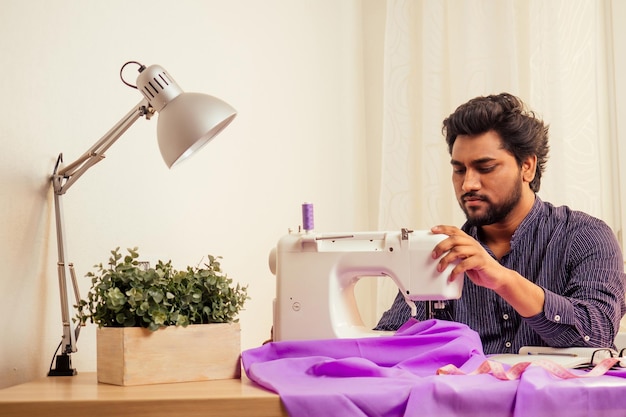 Hübscher indischer Schneidermann In einem stilvollen Hemd, das mit violettem Baumwolltextil in der Heimwerkstatt arbeitet: Auf dem Tisch eine Lampe und ein Topf mit Pflanzen.