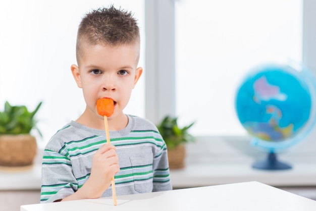 Hübscher Junge, der frische Karotte auf einem Stock zu Hause isst. Das Kind isst Gemüse in seinem Zimmer.