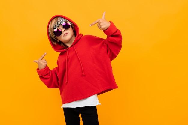 Hübscher Junge mit einem Kopftuch auf seinem Kopf in einem roten Kapuzenpulli mit Brille, die auf isoliertem Orange aufwirft