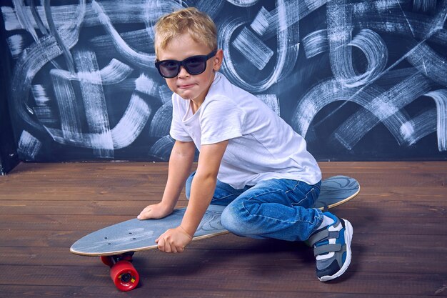 Hübscher Junge mit Sonnenbrille sitzt auf einem großen Skateboard. Kleiner Junge, der auf einem Brett reitet