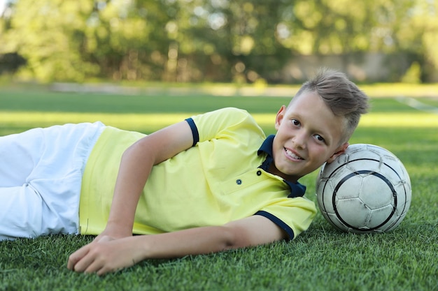 Hübscher Jungenfußballspieler liegt mit dem Ball auf dem Fußballplatz