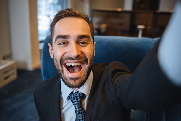 Hübscher junger aufgeregter Geschäftsmann, der Anzug sitzt, der an der Hotellobby sitzt, Handy benutzt und ein Selfie macht