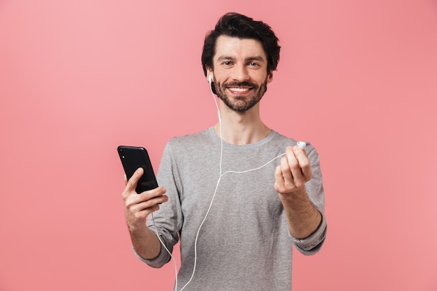 Hübscher junger bärtiger brünetter Mann, der Pullover trägt, der über Rosa steht, Musik mit Kopfhörern hörend, Handy haltend