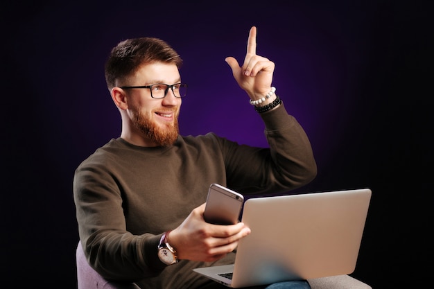 Foto hübscher junger bärtiger freiberufler in freizeitkleidung und brille, der an seinem arbeitsplatz sitzt und auf dem handy spricht und seinen zeigefinger hebt.