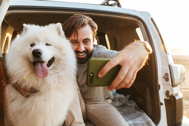 Hübscher junger bärtiger Mann, der hinten in seinem Auto sitzt, mit Hund am Strand spielt und ein Selfie macht