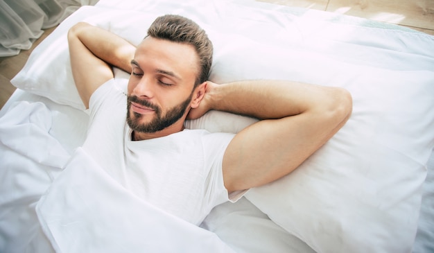 Hübscher junger bärtiger Mann schläft in einem weißen großen Bett in einem bequemen Schlafzimmer