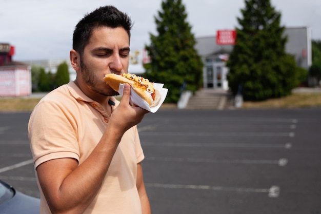 Hübscher junger brünetter Mann, der Hot Dog auf dem Parkplatz nahe der Tankstelle isst.