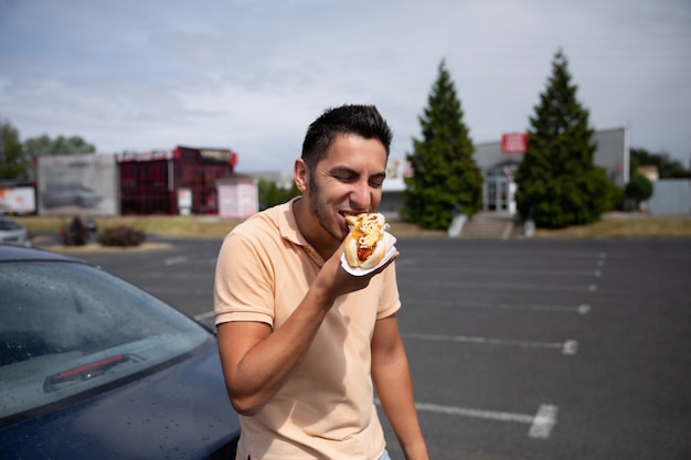 Hübscher junger brünetter Mann, der Hot Dog auf dem Parkplatz nahe der Tankstelle isst.