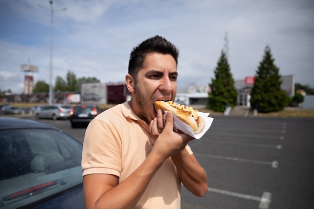 Hübscher junger Brunettemann, der Hotdog im Parkplatz nahe der Tankstelle isst.