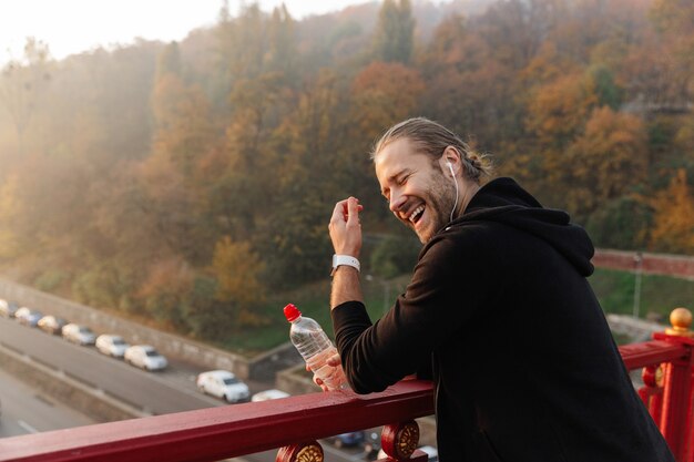 Hübscher junger fitter Sportler, der Musik mit drahtlosen Kopfhörern auf einer Brücke hört, Wasserflasche hält und lacht