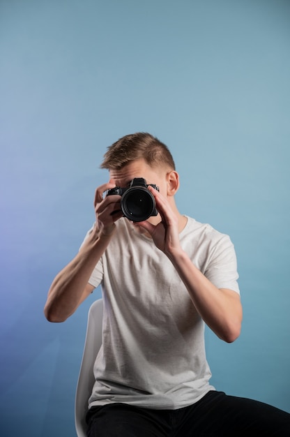 Foto hübscher junger fotograf, der kamera auf blauem hintergrund verwendet. mann, der eine professionelle kamera verwendet. porträt des kerls, der fotokamera betrachtet