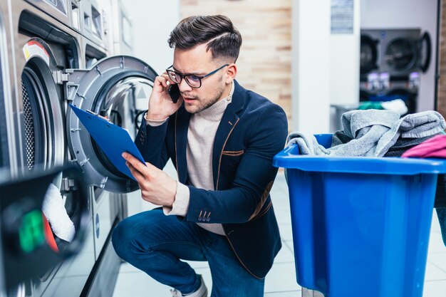 Hübscher junger Geschäftsmann, der am Handy spricht, während er seine wöchentliche Wäsche in einem Waschsalon tut.