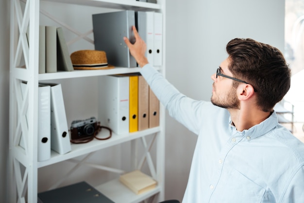 Hübscher junger Geschäftsmann mit Brille, der Ordner aus dem Regal im Büro nimmt