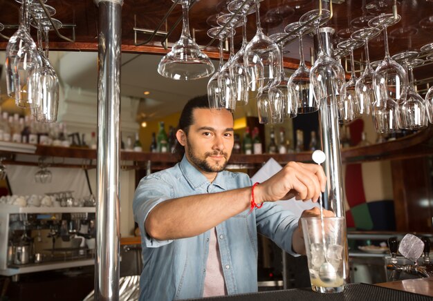 Hübscher junger lächelnder Barmixer, der Stahlsprudellöffel im Glas mit Eis hält.