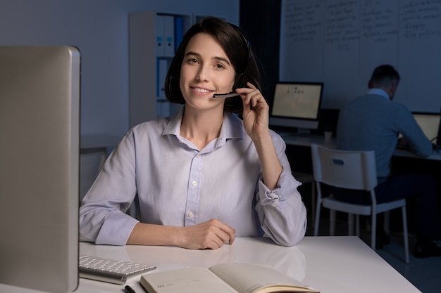 Hübscher junger lächelnder Kundendienstmitarbeiter mit Headset, der mit Kunden online im Büro spricht