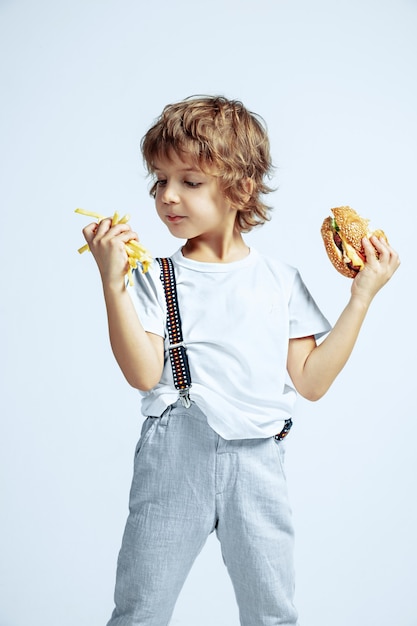 Hübscher junger lockiger Junge in Freizeitkleidung auf weißer Wand. Burger mit Bratkartoffel essen. Kaukasischer männlicher Vorschulkind mit hellen Gesichtsgefühlen. Kindheit, Ausdruck, Spaß, Fast Food.