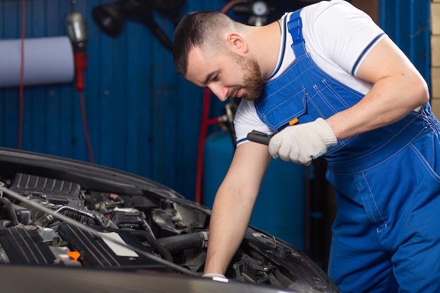 Hübscher junger männlicher Automechaniker in der speziellen Uniformkleidung, die eine Taschenlampe hält, auf der Suche nach Panne und Reparatur unter der Motorhaube im Automotor in einer Autowerkstatt