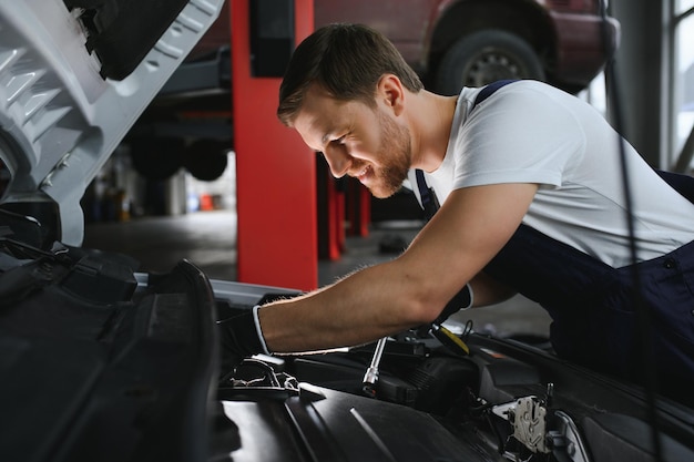 Foto hübscher junger männlicher automechaniker in spezieller einheitlicher kleidung, der eine taschenlampe hält, die nach pannen sucht und unter der motorhaube im automotor in einer autowerkstatt repariert