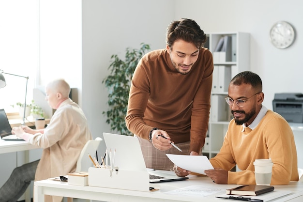 Hübscher junger Manager, der auf Papier zeigt, während er mit einem Kollegen im Büro über die Projektstrategie diskutiert