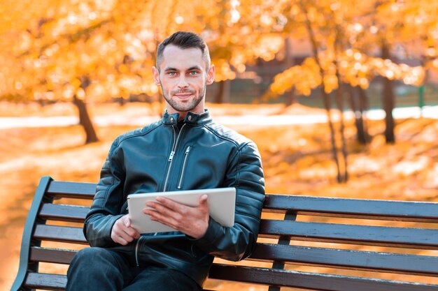 Hübscher junger Mann, der auf der Bank sitzt und großen weißen Tablet-PC im schönen Herbstpark verwendet