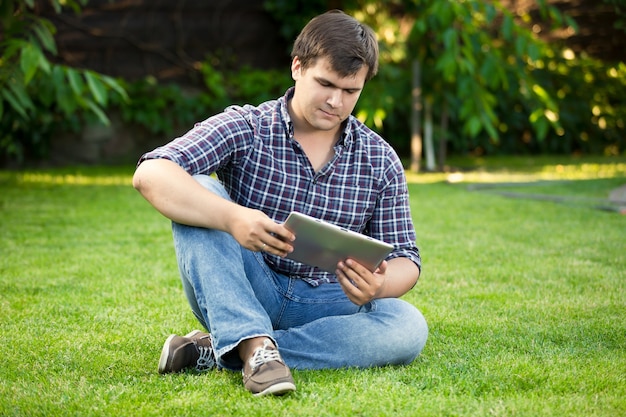 Hübscher junger Mann, der auf Gras im Park sitzt und digitales Tablet benutzt