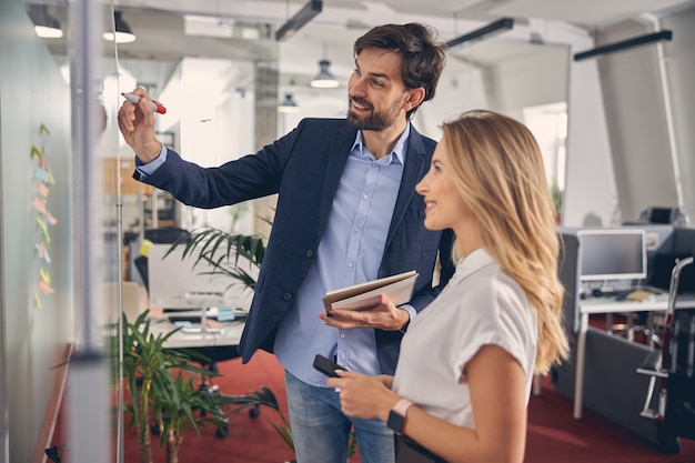 Hübscher junger Mann, der auf Whiteboard schreibt und lächelt, während er neben charmanter blonder Dame steht