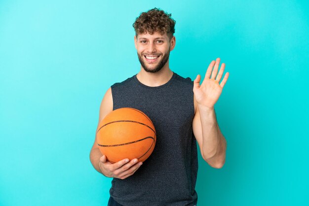 Hübscher junger Mann, der Basketball isoliert auf blauem Hintergrund spielt und mit der Hand mit glücklichem Ausdruck grüßt